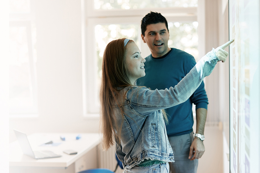 Middle school age girl touches interactive screen with teacher looking on
