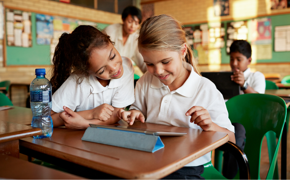 Two smiling elementary age girls using a tablet while sharing a school desk